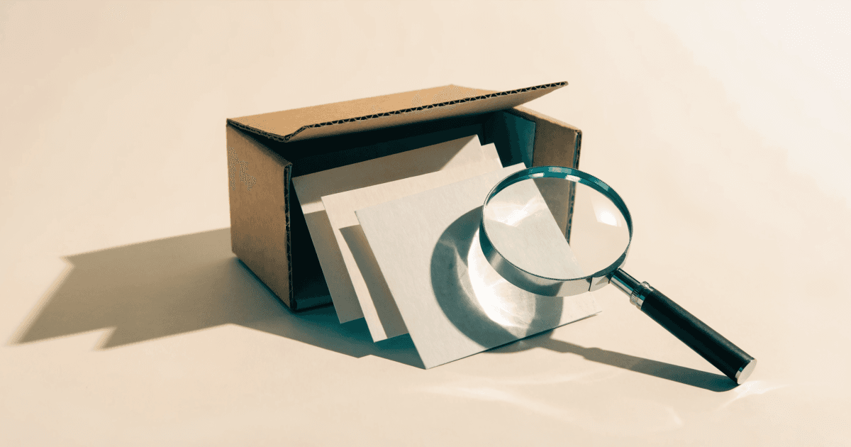 An open cardboard shipping box sits on a cream surface with three blank white cards fanning out from it, accompanied by a magnifying glass examining the documents.