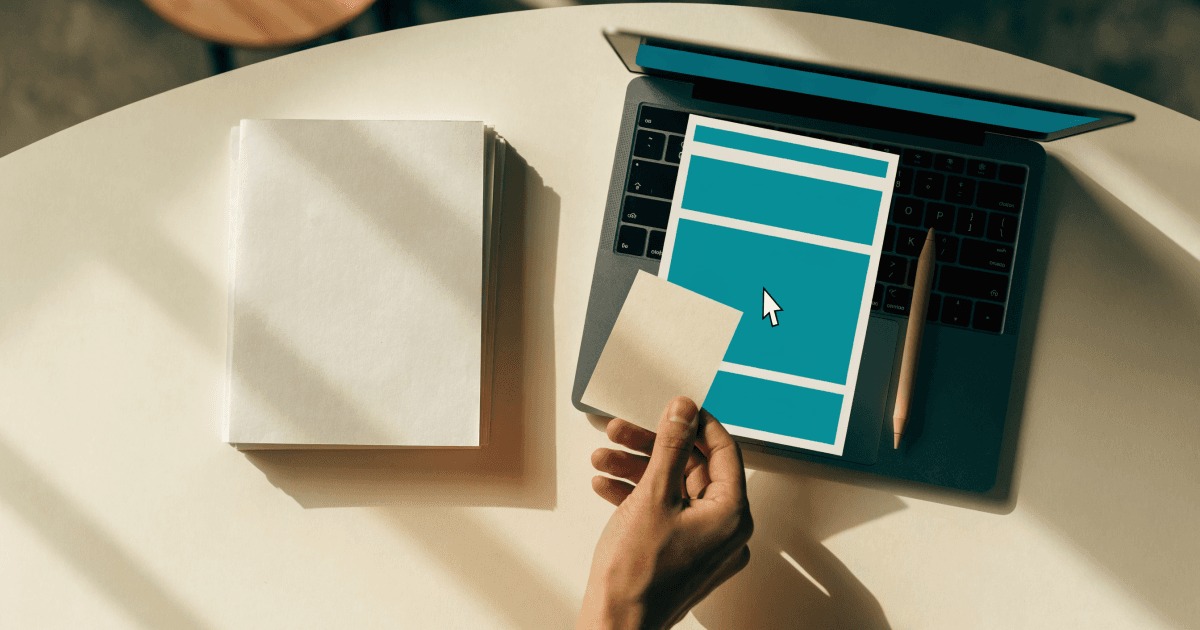 Overhead view of a hand holding a paper card over a laptop displaying a teal document layout on a cream desk.