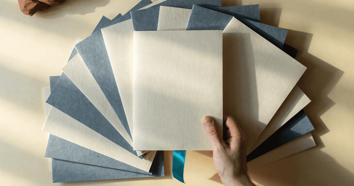 A hand selects a blank cream document folder with a teal ribbon from a fanned stack of neutral folders on a warm desk.