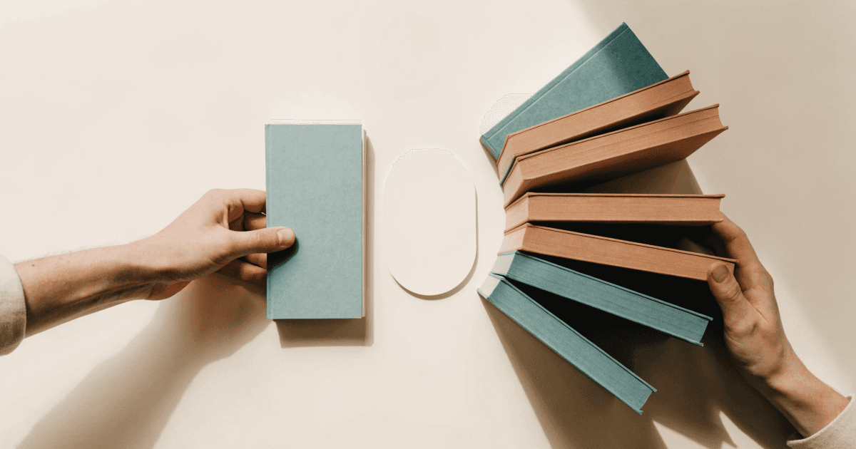 Top-down view of a hand holding a single slim book opposite a hand fanning six thick books around a central blank card on a cream desk.