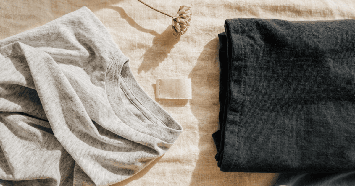 A flat-lay comparison of a lightweight heather-gray t-shirt and a heavyweight charcoal t-shirt on a cream linen background with a blank care label and dried cotton flower.