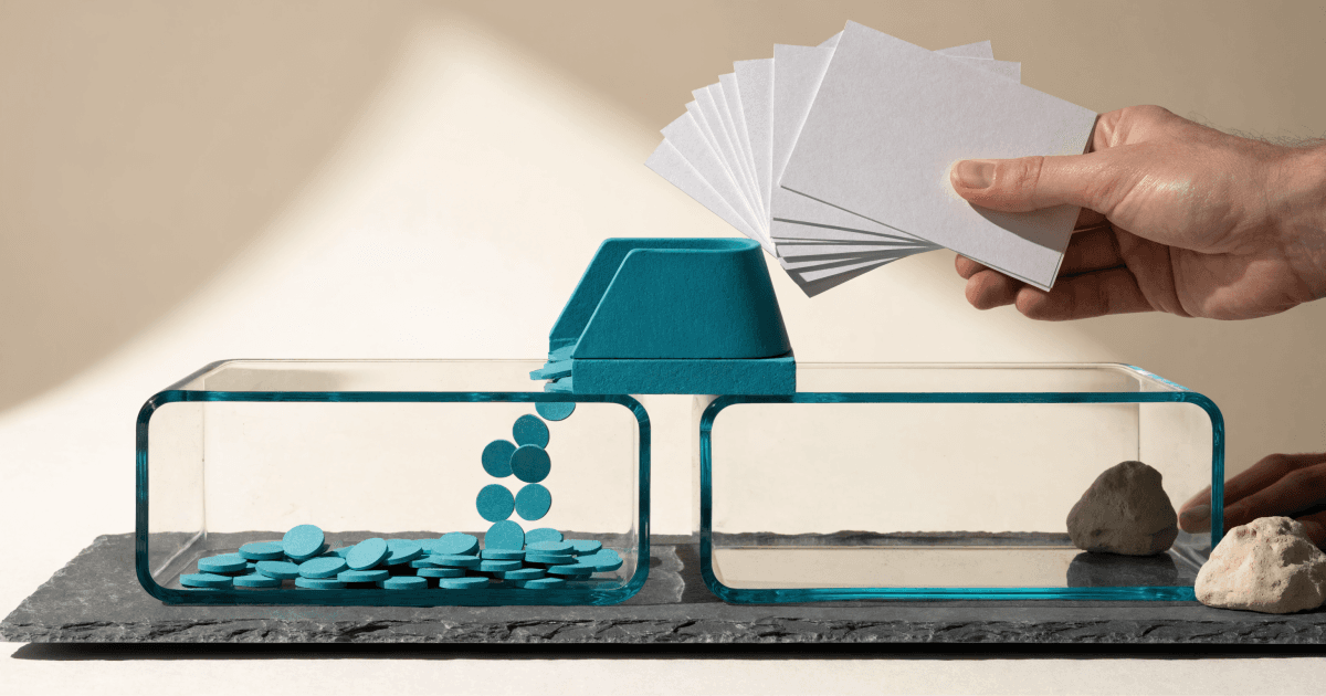 A hand feeds blank cards into a teal measuring gate that drops teal tokens into a glass tray, while an adjacent empty tray holds stones.